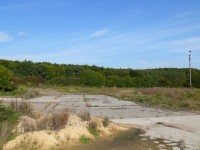 Hier eine Ansicht ähnlich des Werksfotos. Links über den Kanthölzern ist das alte Fundament der Halle zu erkennen. Darüber sind leicht &quot;rötliche&quot; Büsche, links davon ist das Tor im Zaun. Rechts ist die L141 zu sehen, links davon stand damals das eingezäunte Haus. Die HLS muss ungefähr da gestanden haben, wo die Balken zu Ende sind, etwas weiter oben wo der hellgrüne Busch anfängt.